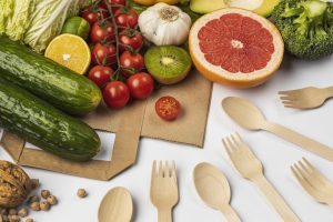 Assortment of fresh produce—cucumbers, tomatoes, citrus, leafy greens—arranged on a table with wooden utensils and plates.